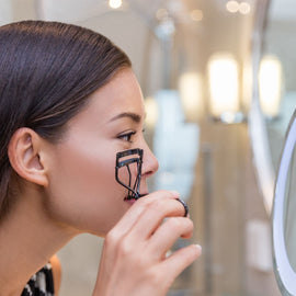 An Asian woman carefully curling her eyelashes in front of a mirror, focusing on her reflection with a gentle expression.